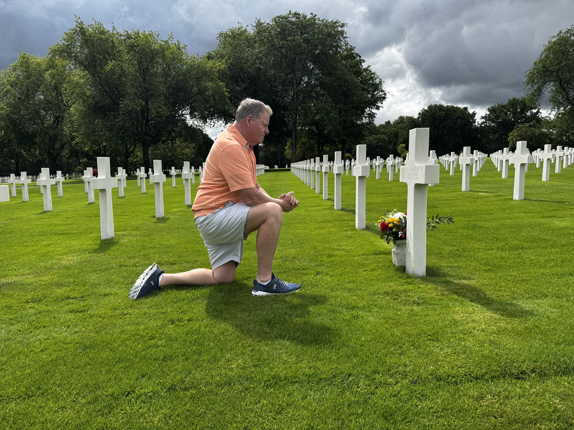 Tom Breen kneeling at the grave of his father's best friend at Lorraine American Cemetery, on a 42nd Rainbow Division battlefield tour in his father's footsteps with historian Erin Faith Allen